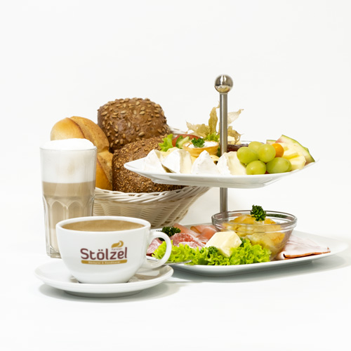 Frühstück mit Brötchenkorb, einem Latte Macchiato, einem Kaffee mit dem Bäckerei Stölzel Logo sowie einer Etagere mit Obst & Belag, freigestellt vor weißem Hintergrund