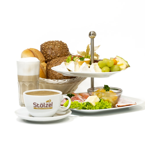 Frühstück mit Brötchenkorb, einem Latte Macchiato, einem Kaffee mit dem Bäckerei Stölzel Logo sowie einer Etagere mit Obst & Belag, freigestellt vor weißem Hintergrund